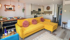 A modern living room with a yellow sectional sofa, a dining table, and a kitchen area. - 2 De Courcey House (Dartmouth)