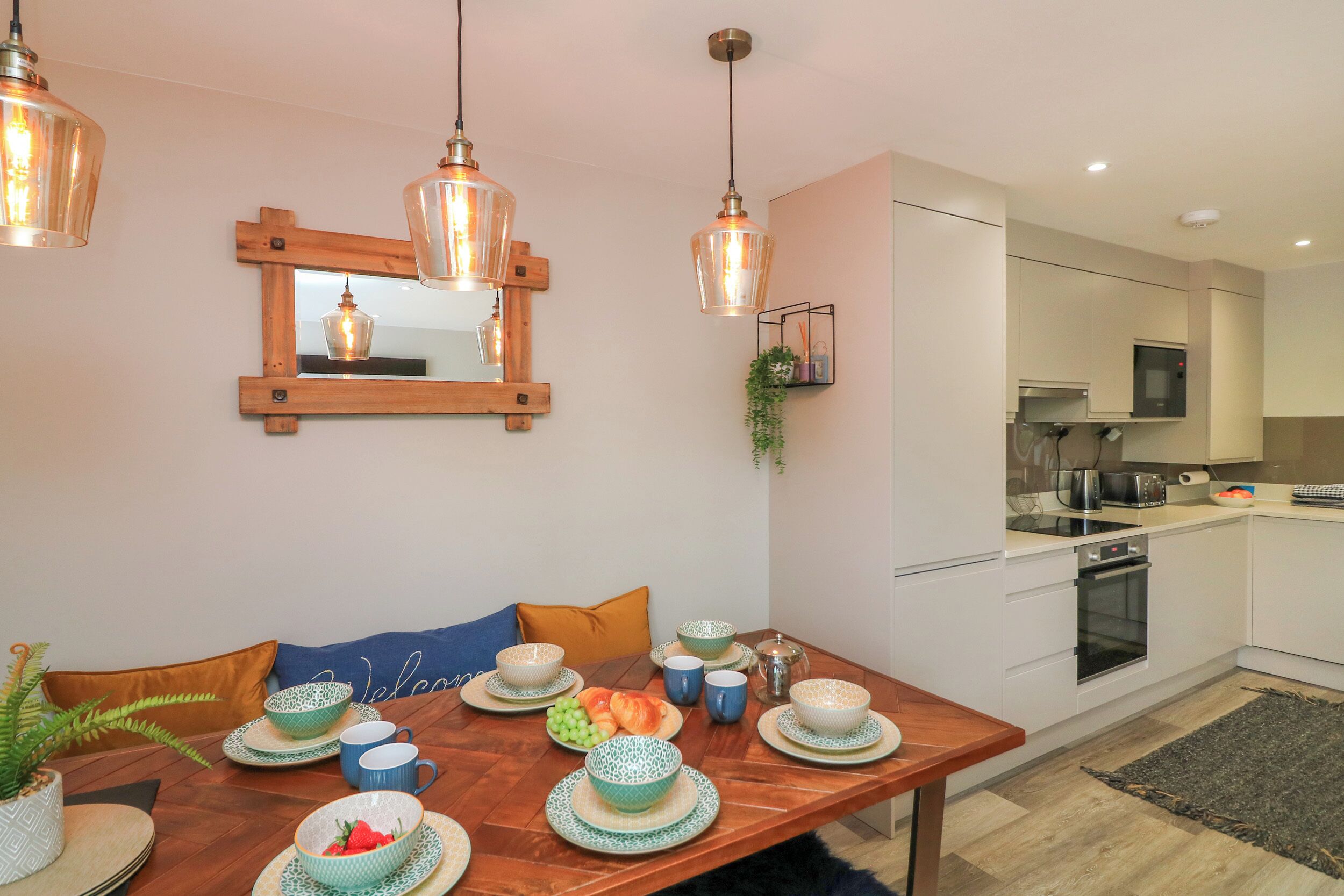 A modern kitchen with white cabinets, a microwave, and an oven. There is a dining table with blue and green patterned plates and cups.