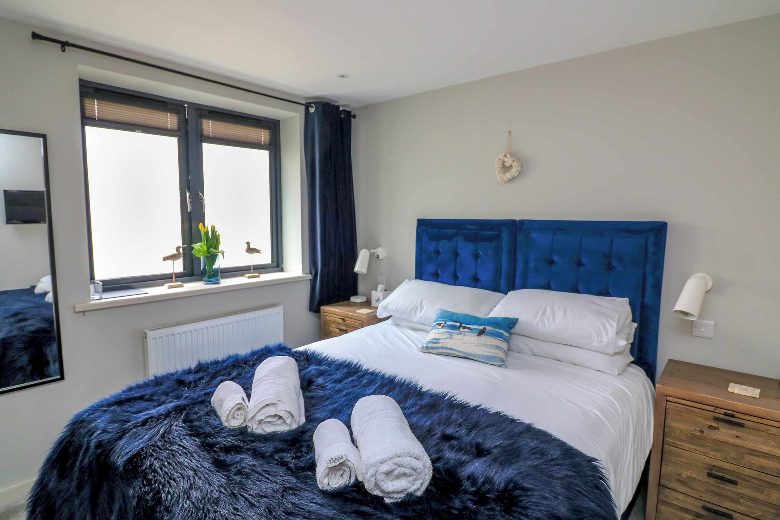 Un dormitorio con una cama grande, cabecera azul, ventana con cortinas, mesita de noche con lámpara, y una lámpara de pared.