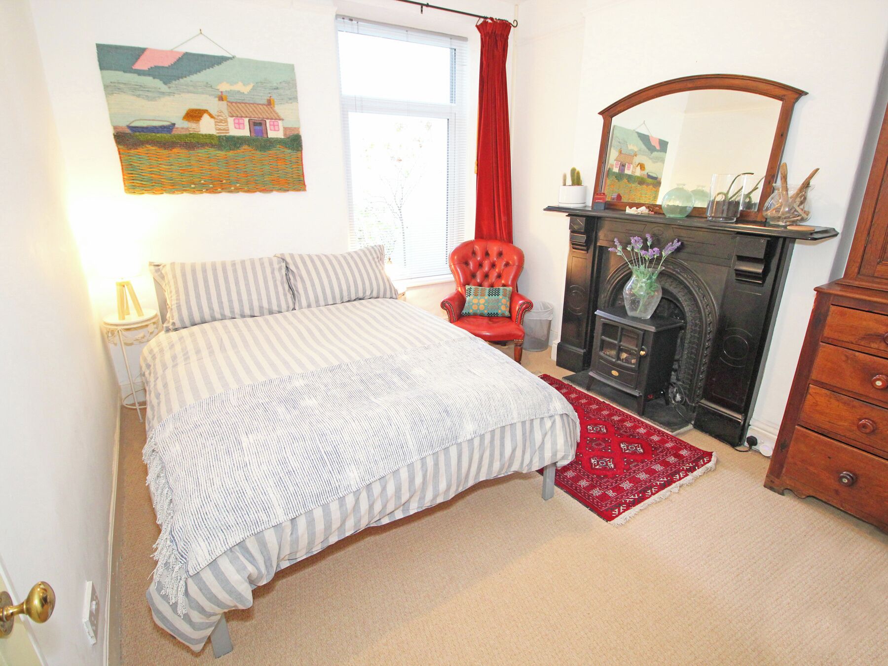 A bedroom with a bed, a red chair, a fireplace, and a mirror.
