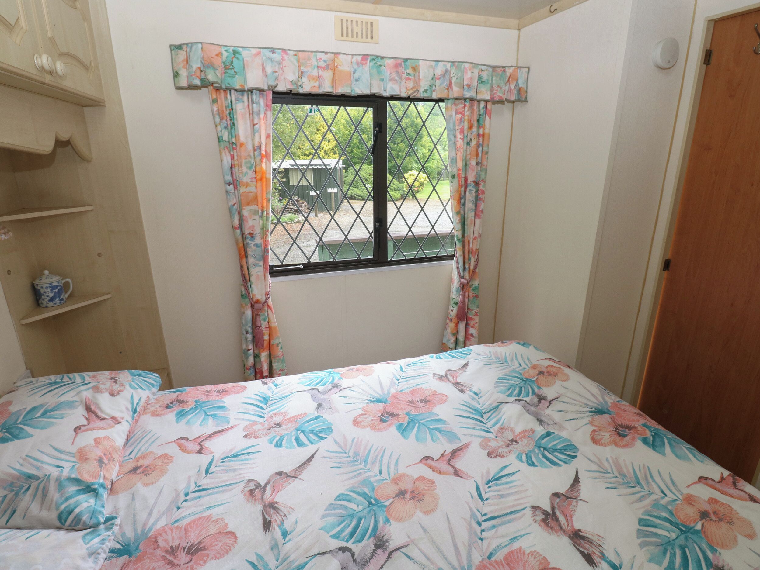 Ein Schlafzimmer mit Bett, einem Fenster mit floralem Vorhang, einer Holzablage mit Teekanne und einer Tür.