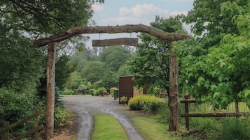 A rustic wooden archway leading to a gravel path surrounded by lush greenery and a wooden structure in the distance.