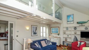 A spacious living room with a blue sofa, a red armchair, and a wooden coffee table. There are two windows with curtains, a glass railing on the upper level, and a bookshelf with decorative items. - Seapie Corner (Yarmouth)