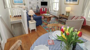 A well-lit living room with a blue sofa, a red armchair, a wooden coffee table, and a dining table set for two. There are bookshelves, a television, and a window with curtains. - Seapie Corner (Yarmouth)