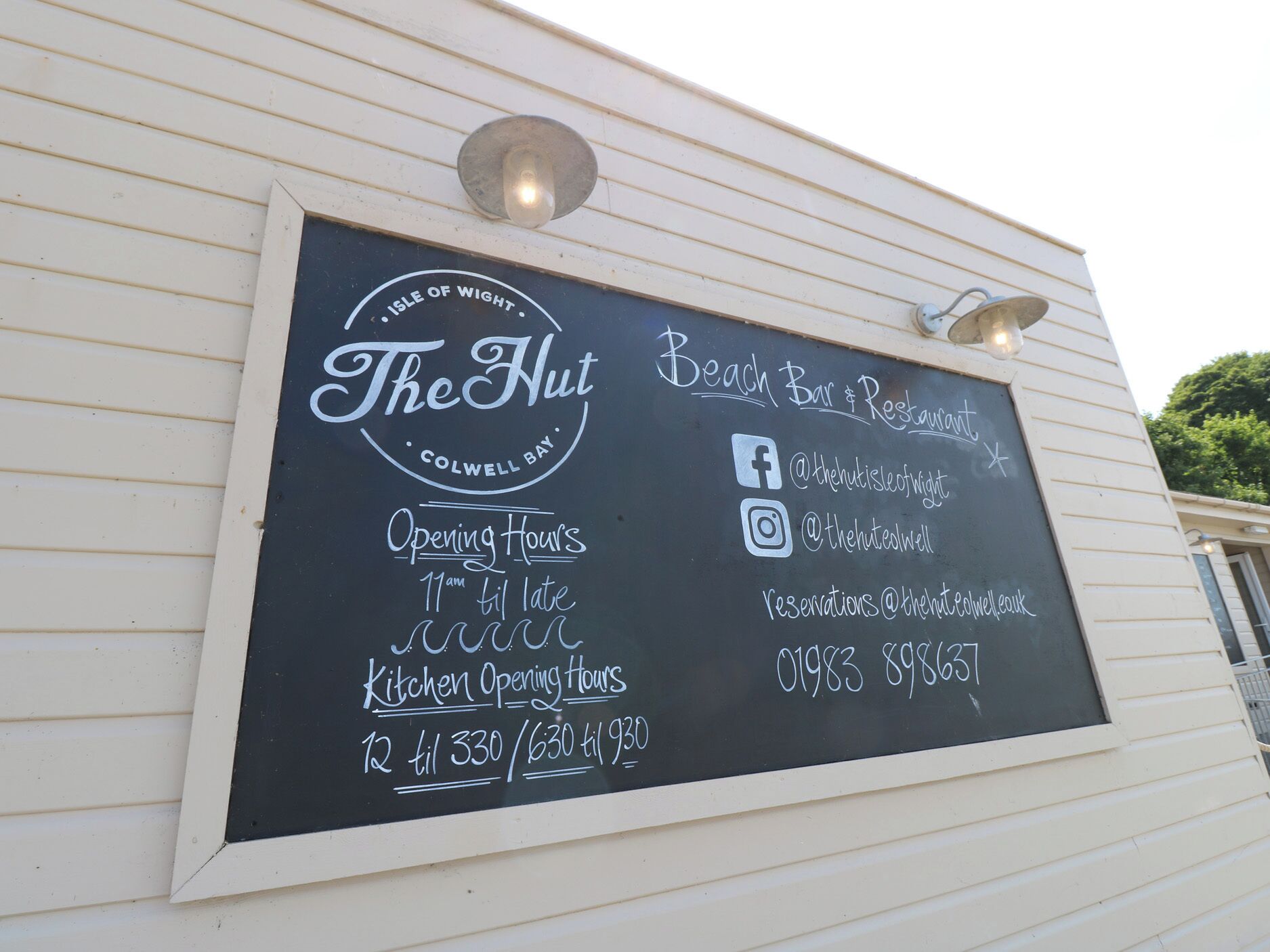 A chalkboard sign for 'The Hut' with opening hours, Facebook and Instagram handles, and a phone number.