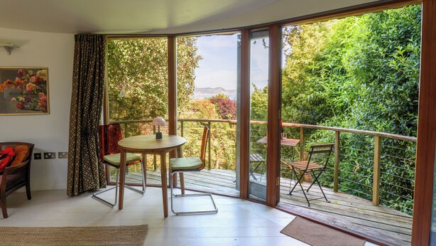 A room with a sliding glass door leading to a balcony with a view of greenery and a distant landscape. - The Bothy (Lyme Regis)