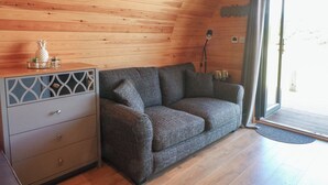 A cozy room with a wooden ceiling, a grey sofa, a small cabinet with drawers, and a door leading outside. - Paradise (Market Drayton)