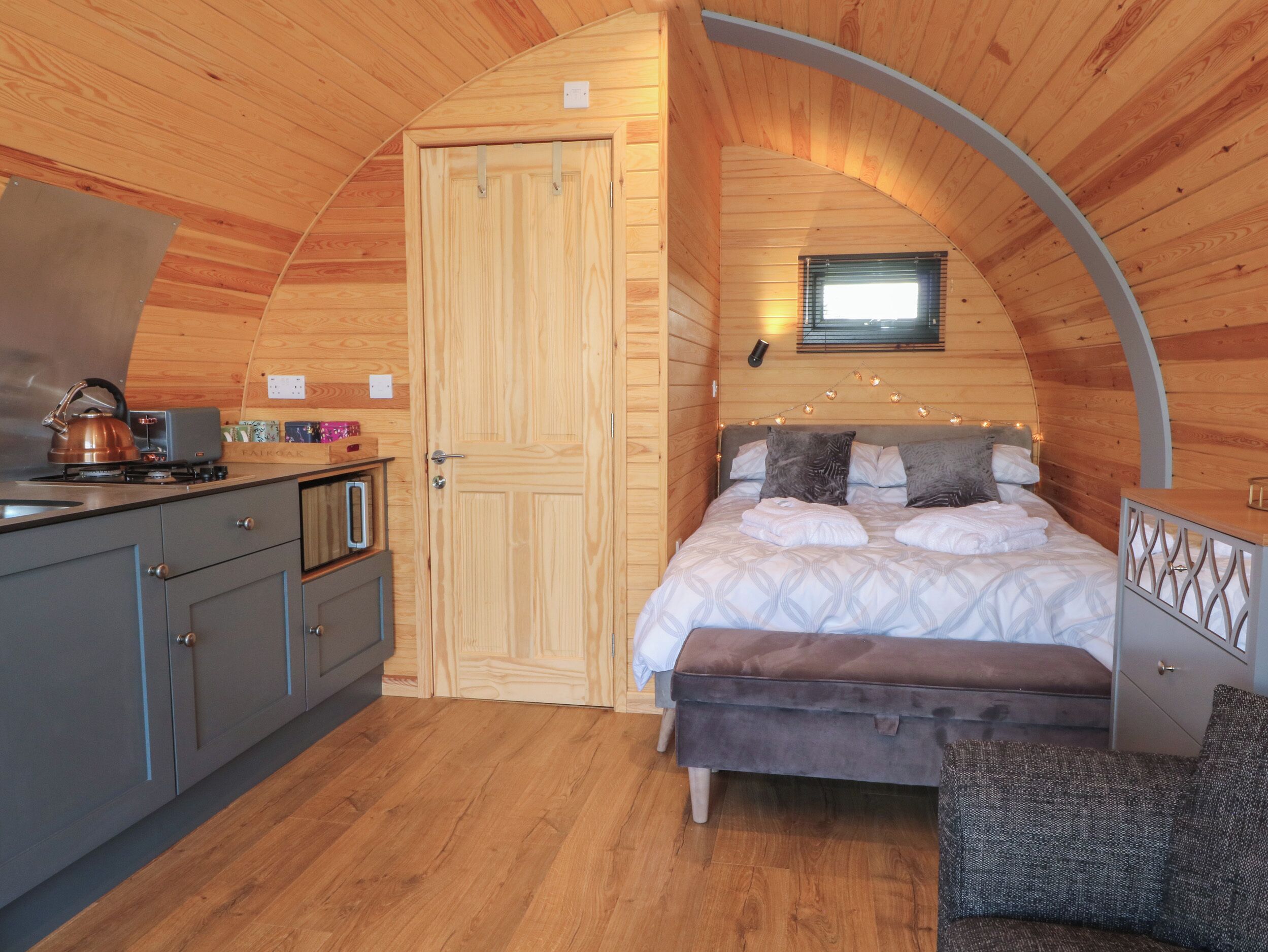 A cozy, wooden cabin-style room with a curved ceiling, a bed with pillows, a bench, a small table, and a kitchenette with a kettle.