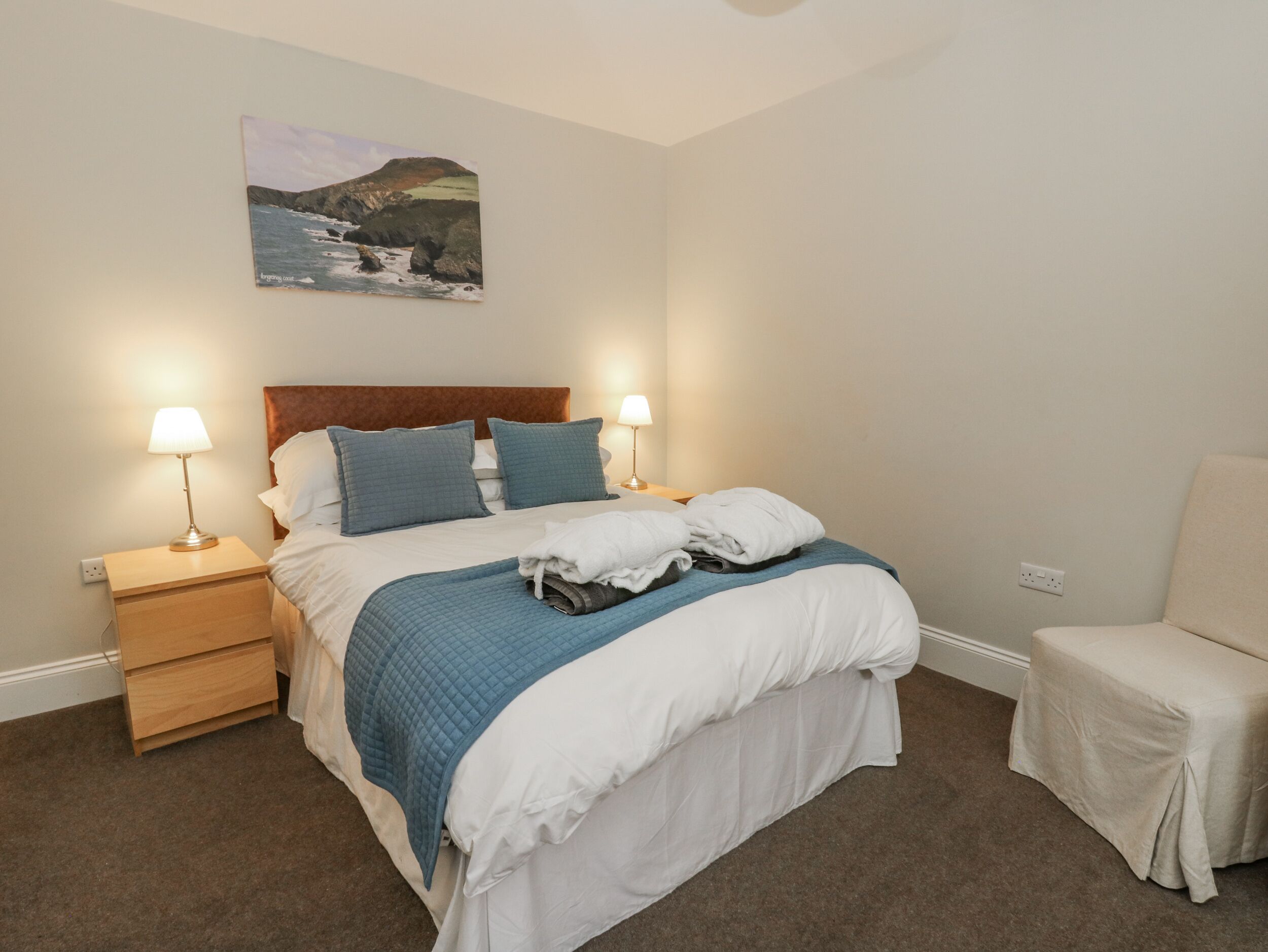 A neatly made bed with a blue quilt, two bedside lamps, a nightstand, and a small chair.