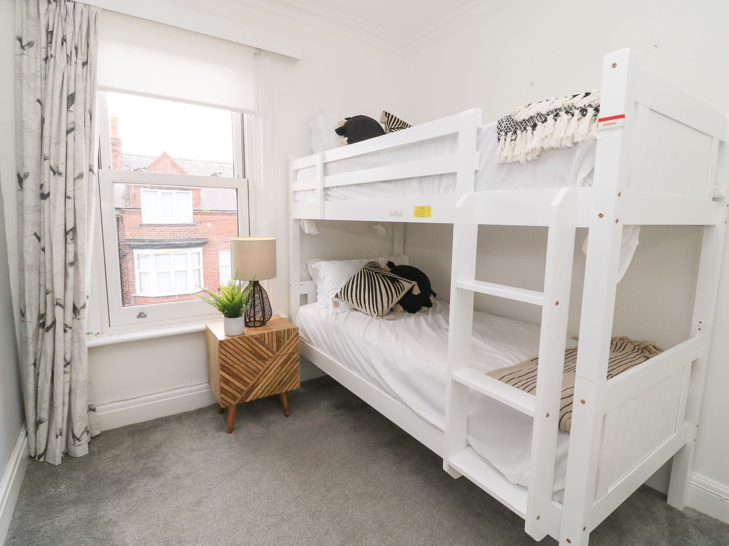 A bunk bed room with a window, a small wooden side table with a lamp, and a plant.