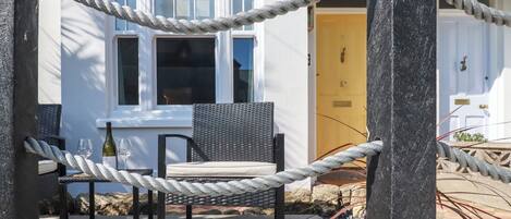 A wooden deck with a wicker chair and a table, a rope barrier, and a white building with a yellow door in the background.