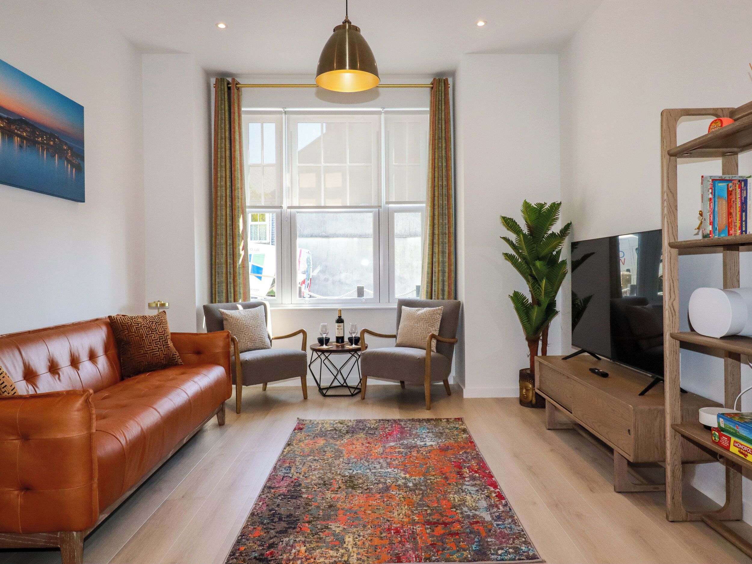 A modern living room with a brown leather sofa, two armchairs, a coffee table, a colorful rug, a television, and a bookshelf.