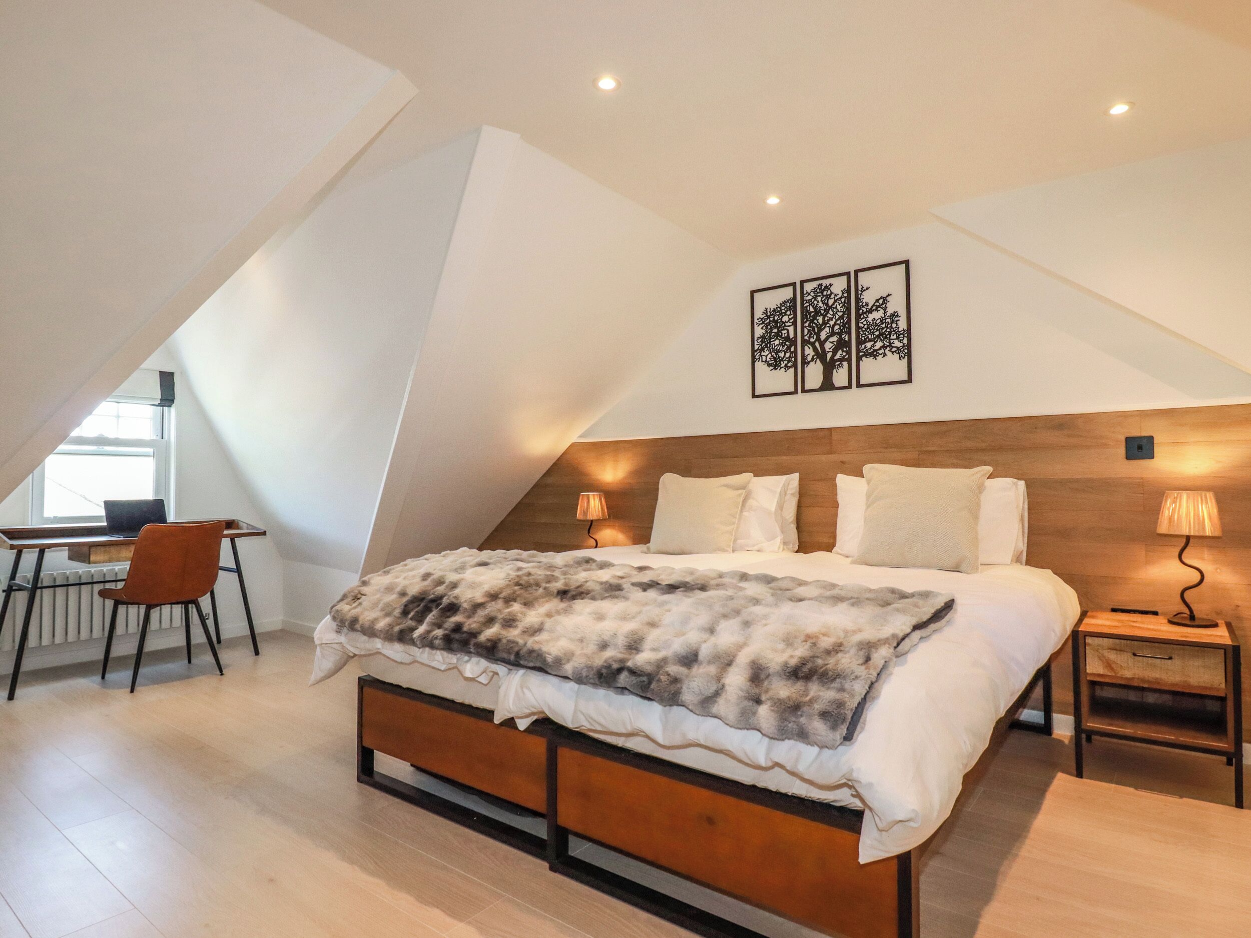 Un dormitorio con techo inclinado, una cama con manta mullida, un escritorio con silla y lámparas fijadas a la pared.