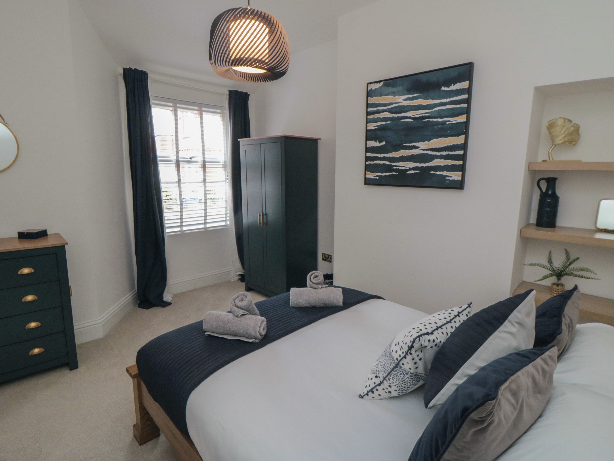 Un dormitorio con cama, armario, cómoda, ventana con cortinas, lámpara colgante y una obra de arte en la pared.