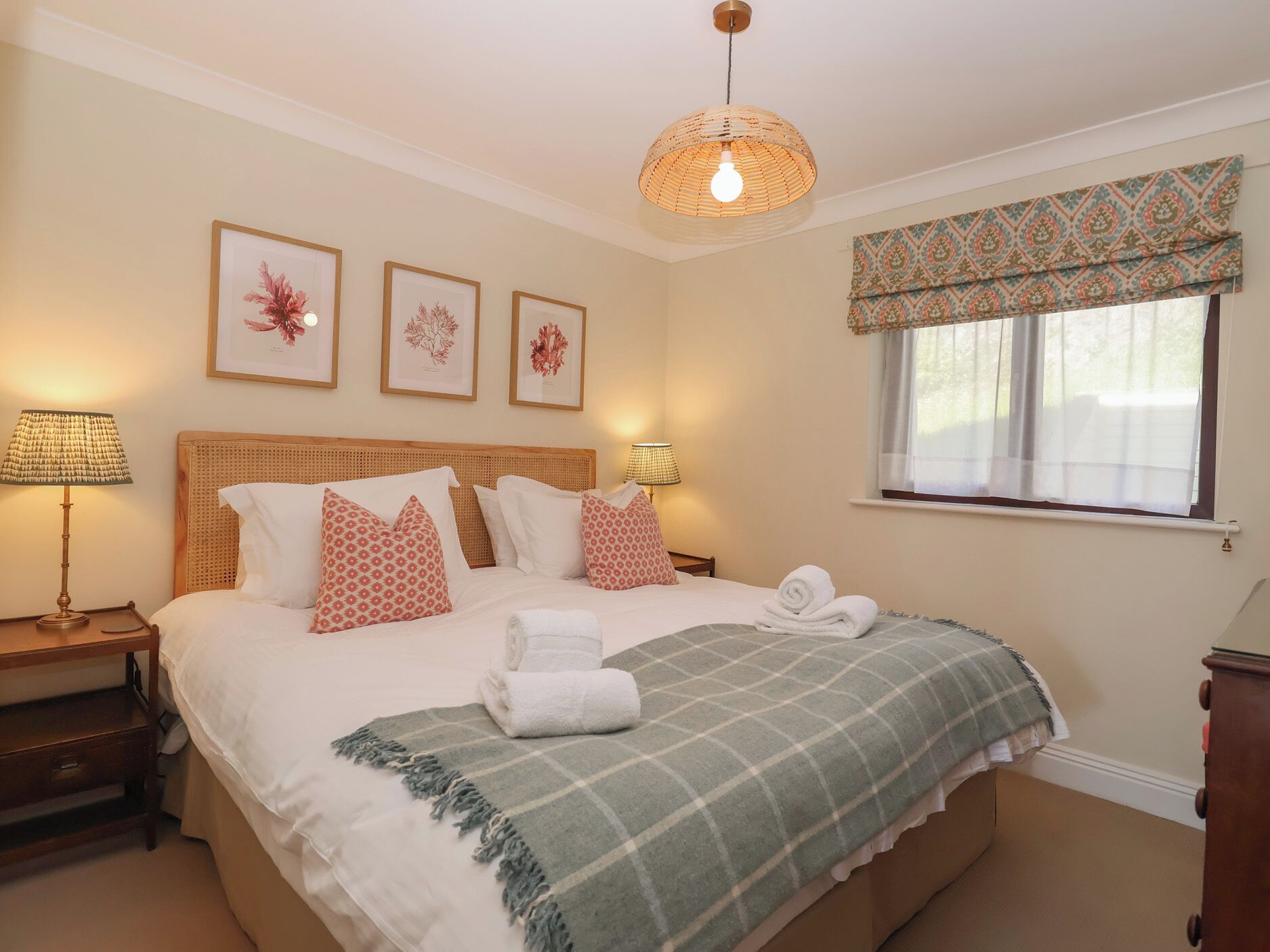 A neatly arranged bedroom with a bed, bedside lamps, a window with a valance, and framed botanical prints on the wall.