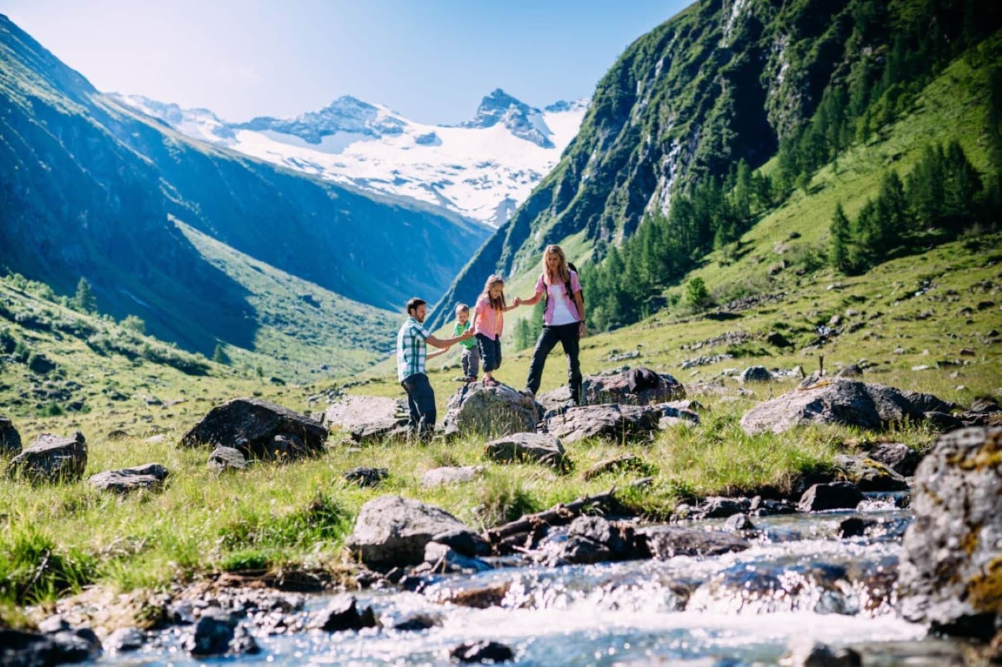 Une famille fait une randonnée dans une région montagneuse, avec un ruisseau et des sommets enneigés en arrière-plan.