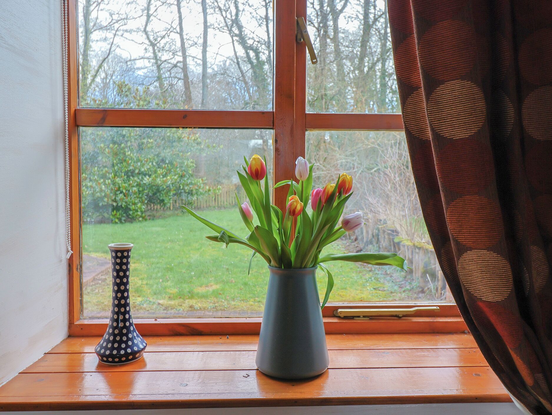 A window with a view of a garden, a vase with tulips, and a polka-dotted vase on the windowsill.