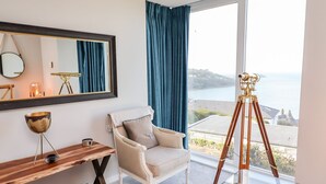 Ett rum med ett stort fönster som ger utsikt över havet, ett teleskop monterat på ett stativ, en stol, ett litet bord i trä och en spegel på väggen.