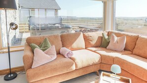 A cozy living room with a large window, a sectional sofa, a floor lamp, and a view of a house and landscape. - 3 Trem y Mor (Holyhead)