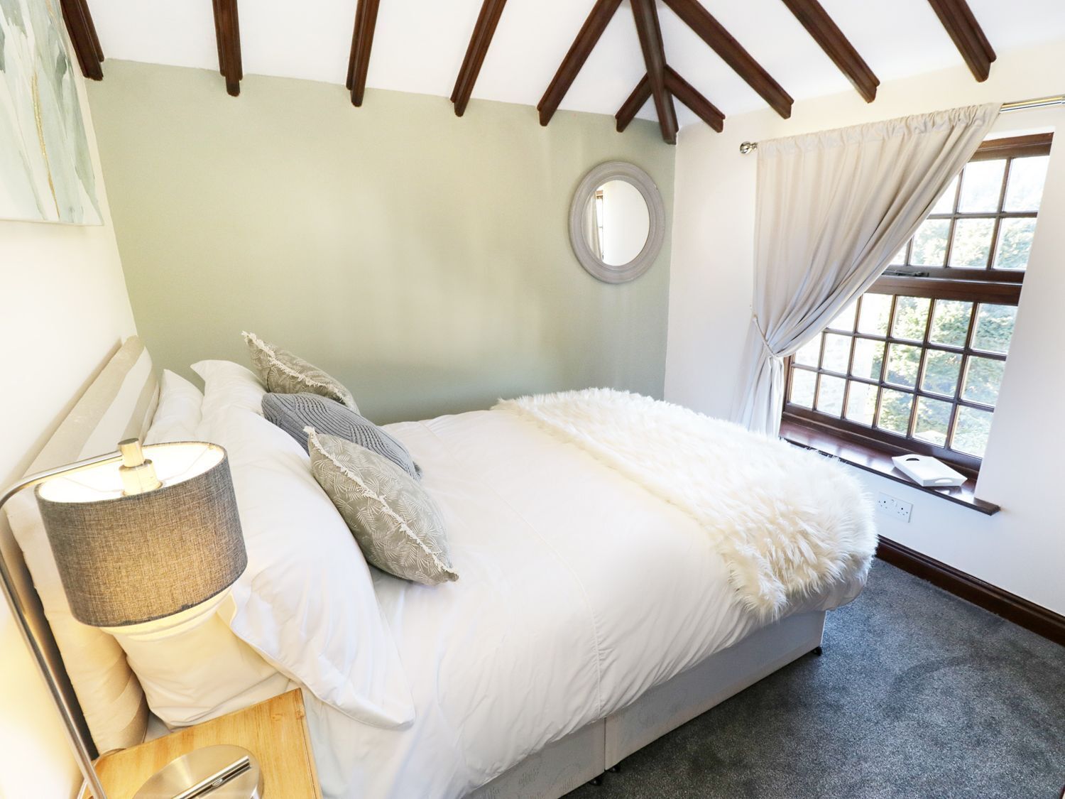 A bedroom with a bed, a bedside lamp, a round mirror, and a window with curtains.