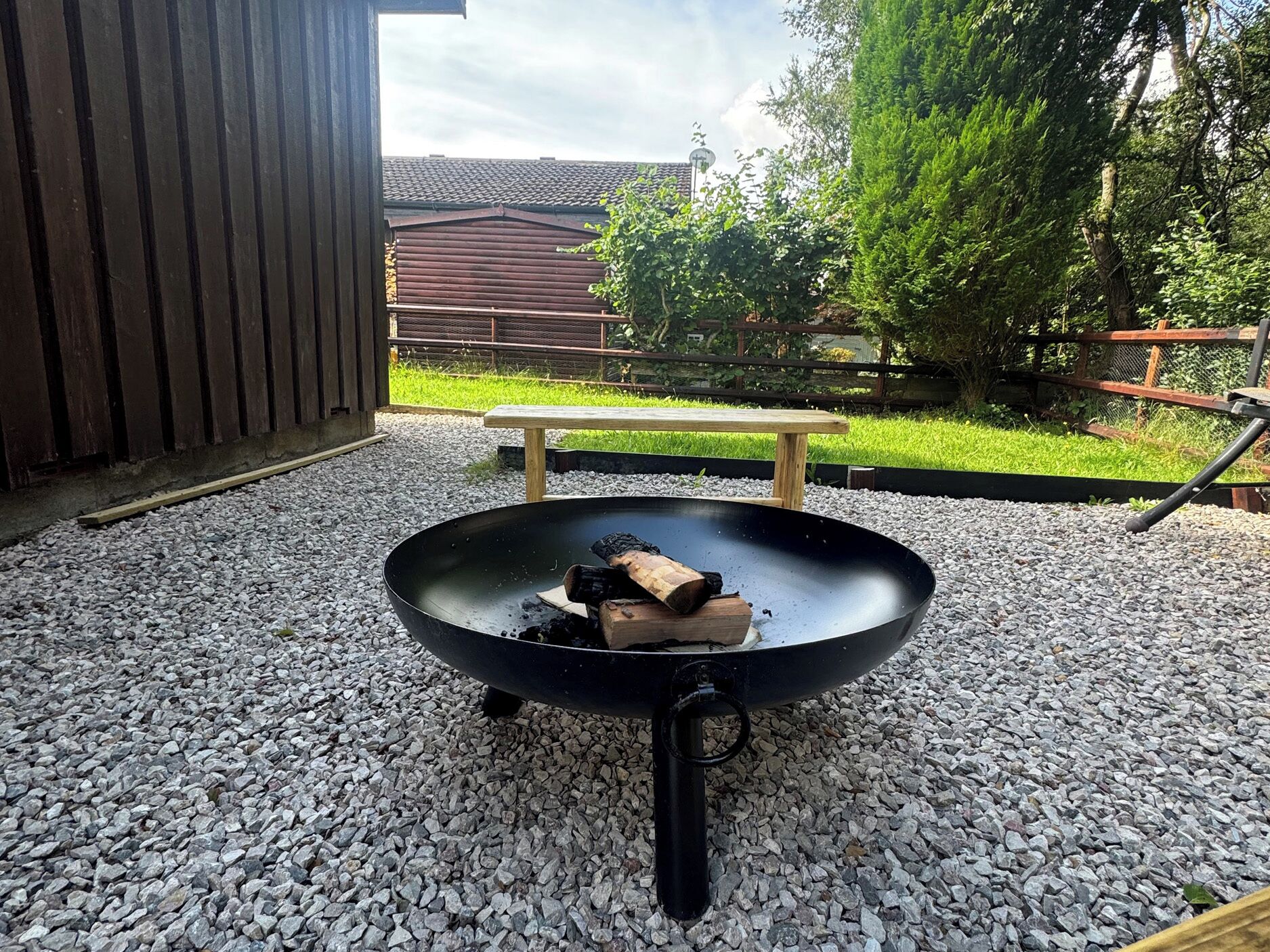 A round outdoor grill on gravel with a bench and a wooden fence in the background.