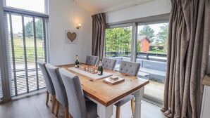 A dining area with a wooden table, grey upholstered chairs, and a view of a green landscape through large windows. - Fig Tree Lodge (Penrith)