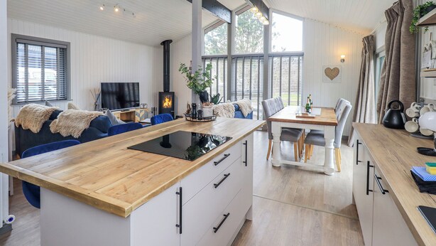 A modern kitchen with a central island, a dining area with a table and chairs, a built-in stove, and large windows. - Fig Tree Lodge (Penrith)