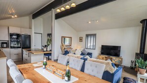 A spacious living area with a wooden dining table, grey upholstered sofas, a flat-screen TV, and a wood-burning stove. - Fig Tree Lodge (Penrith)