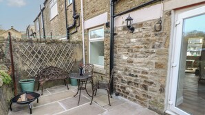 A stone building with a patio area featuring a table and chairs, a potted plant, and a metal fence. - Hollywood Cottage (Barnard Castle)