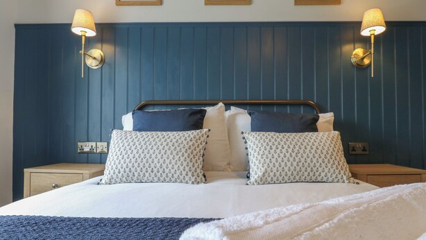 A neatly made bed with patterned pillows, a blue headboard, and wall-mounted lamps. - Hollywood Cottage (Barnard Castle)