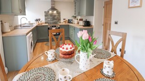 A modern kitchen with light blue cabinets, a stone backsplash, and a wooden dining table set with a cake, tulips, and coffee cups. - Hollywood Cottage (Barnard Castle)