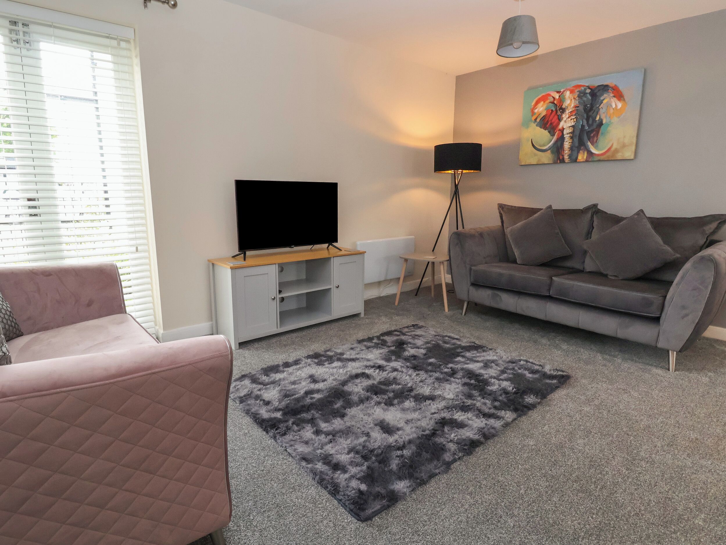 A modern living room with a flat-screen TV, grey sofa, pink armchair, and a painting on the wall.