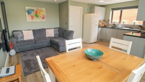 A compact living space with a grey sofa, a wooden dining table with a bowl, a white chair, a small kitchen area with a fridge, and a window with plants. - 6 Fairfax Close (York)