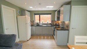 A compact kitchen with a refrigerator, stove, sink, and cabinets. There is a window with a view of a brick wall. A gray sofa is in the foreground. - 6 Fairfax Close (York)
