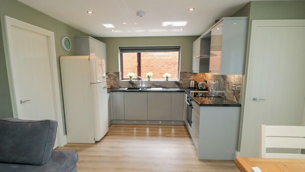 A compact kitchen with a refrigerator, stove, sink, and cabinets. There is a window with a view of a brick wall. A gray sofa is in the foreground. - 6 Fairfax Close (York)