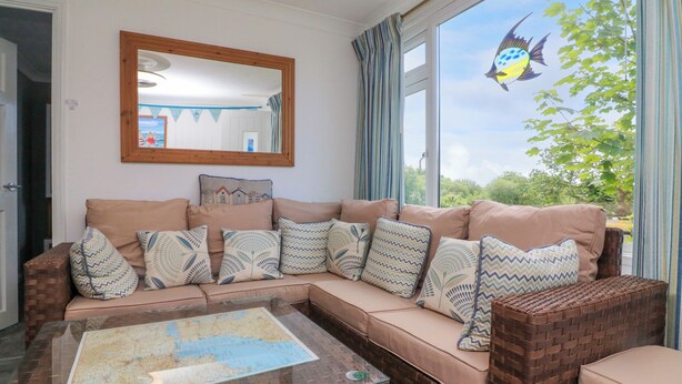 A living room with a sofa, patterned cushions, a glass coffee table with a map, a mirror, and a view of greenery and a fish through the window. - 4 The Park (Bude)