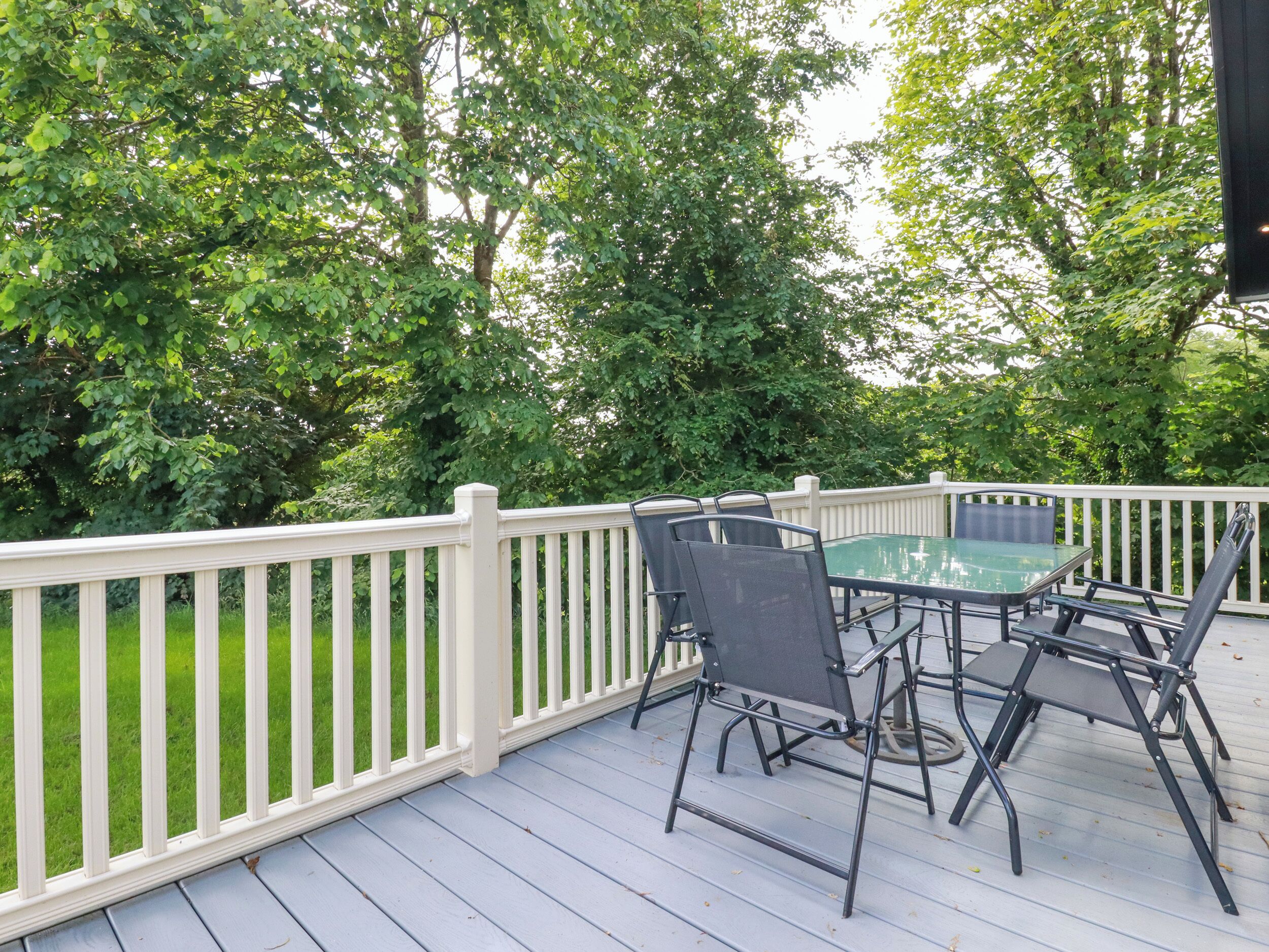 A deck with a glass table and chairs, surrounded by a white railing and lush greenery.
