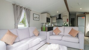 Ein modernes Wohnzimmer mit zwei hellgrauen Sofas, beigen Kissen und einer weißen Tagesdecke. Im Hintergrund ist eine Küche mit Edelstahlgeräten und einem Fenster mit gemusterten Vorhängen zu sehen.