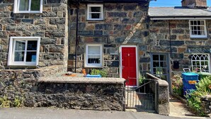 A stone building with a red door, multiple windows, and a small garden area with plants and a blue container. - Riverbank Cottage (Caernarfon)