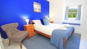A bedroom with a bed, a chair, a nightstand, a lamp, and a window with a view. - Riverbank Cottage (Caernarfon)