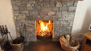 A stone fireplace with a wooden mantel, a fire burning inside, a metal poker, and a basket of firewood. - Riverbank Cottage (Caernarfon)