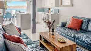 A modern living room with a blue leather sofa, a wooden coffee table, and a view of the kitchen and dining area. - The Cubby Hole (Penzance)