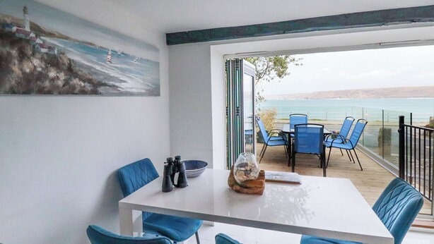 A dining area with a white table and blue chairs, overlooking a scenic view of the sea and a lighthouse. - The Cubby Hole (Penzance)