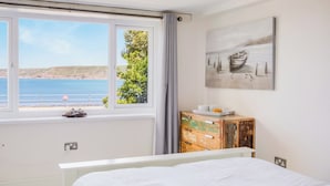 A bedroom with a large bed, a window with a view of the sea, a wooden dresser, and a painting on the wall. - The Cubby Hole (Penzance)