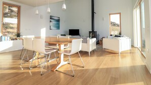 Un vaste espace de vie comprenant une table à manger en bois, des canapés blancs et un éclairage suspendu. Une grande fenêtre offre une vue sur l’extérieur.