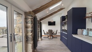 A modern kitchen with dark cabinetry, a dining area with a table and chairs, and a stone wall. - Gaerau Bach (Llanbedr)