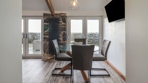 A modern dining area with a glass table, two chairs, a stone wall, and a view through large windows. - Gaerau Bach (Llanbedr)