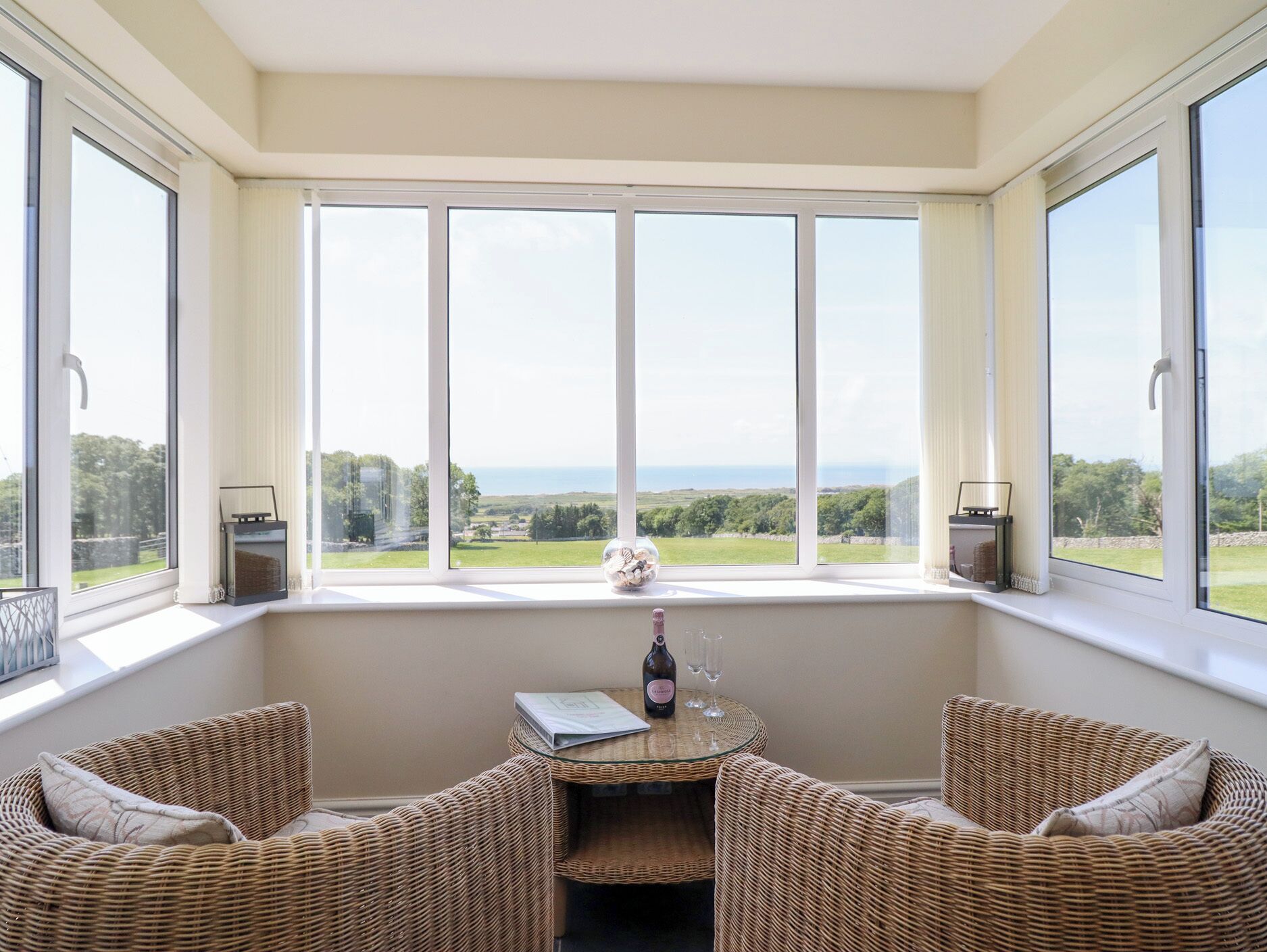 A room with a large window overlooking a scenic landscape, wicker furniture, and a small table with a bottle and glasses.