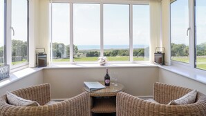 Habitación con un ventanal grande con vistas a un paisaje pintoresco, muebles de mimbre y una pequeña mesa con una botella y copas.