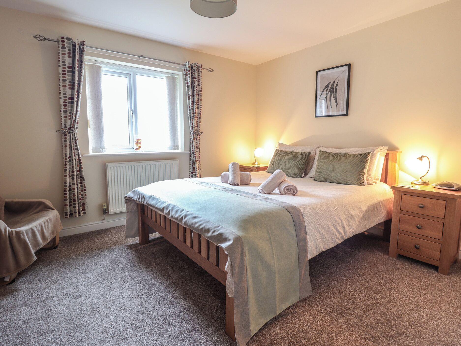 A neatly arranged bedroom with a bed, bedside lamps, a nightstand, a chair, and a window with curtains.